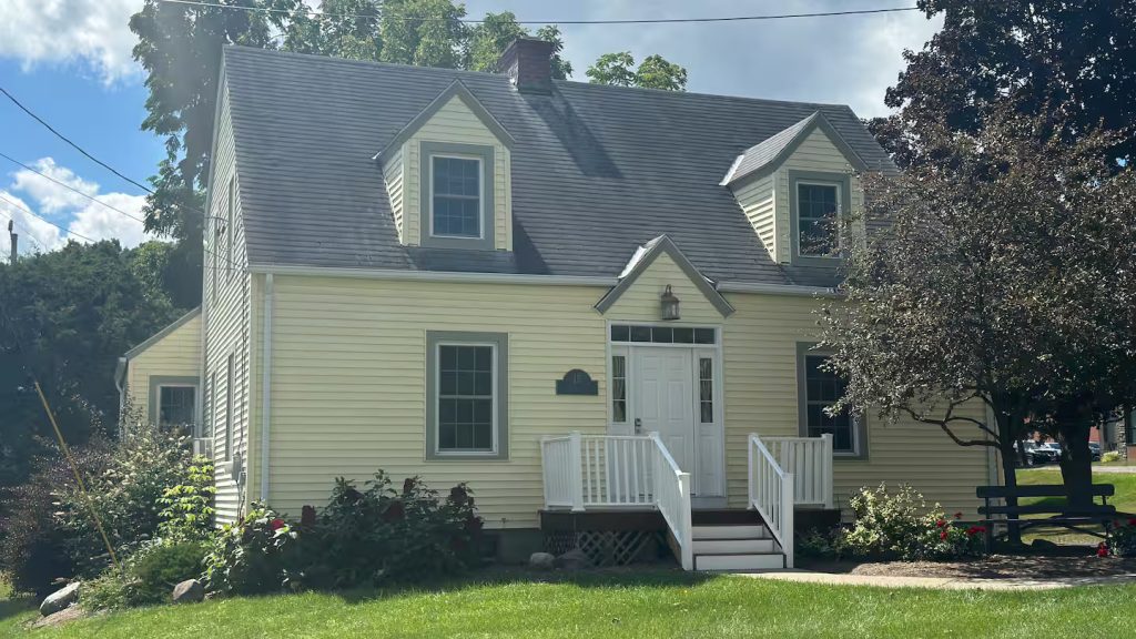 A sunny exterior photo of a light-yellow, two-story house with white trim, a gray shingled roof, dormer windows, front steps and railing, landscaping, and mature trees.